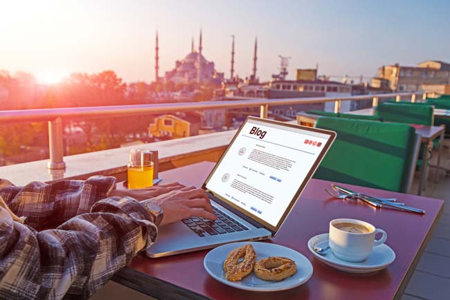 Stemningsbillede fra en terrasse med en person der skriver på en laptop midt i morgenmaden. Smuk solopgang og en stor moske i baggrunden.